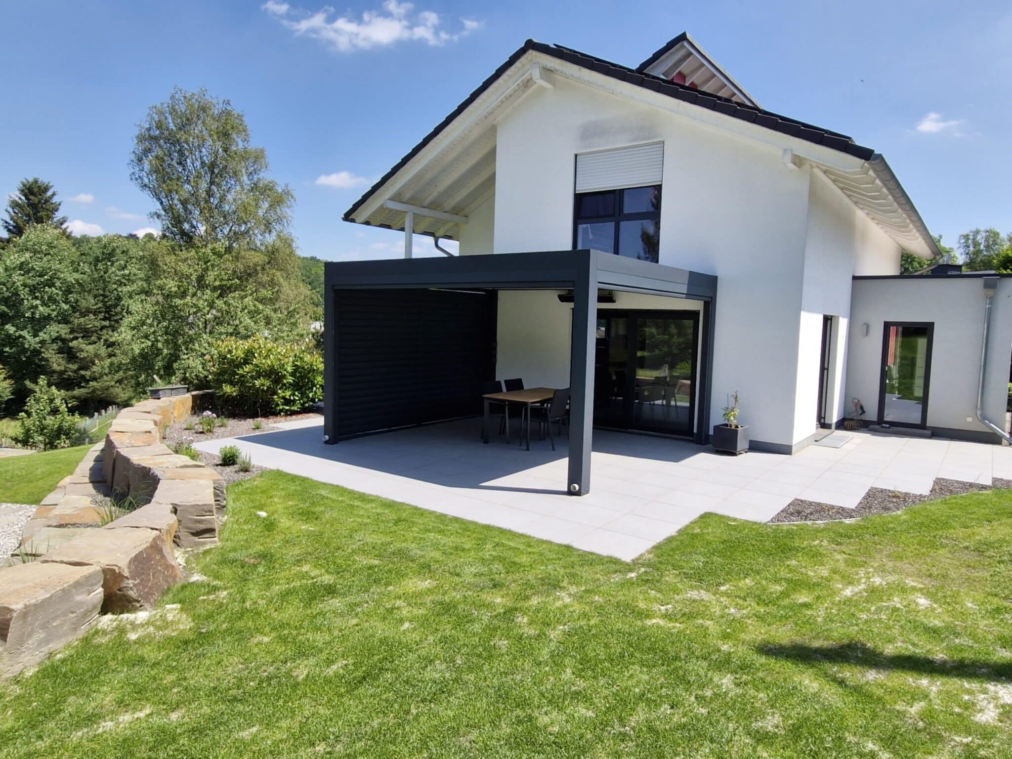 Anthrazitfarbenes Lamellendach mit seitlicher Lamellenwand an einem modernen Wohnhaus bei Sonne