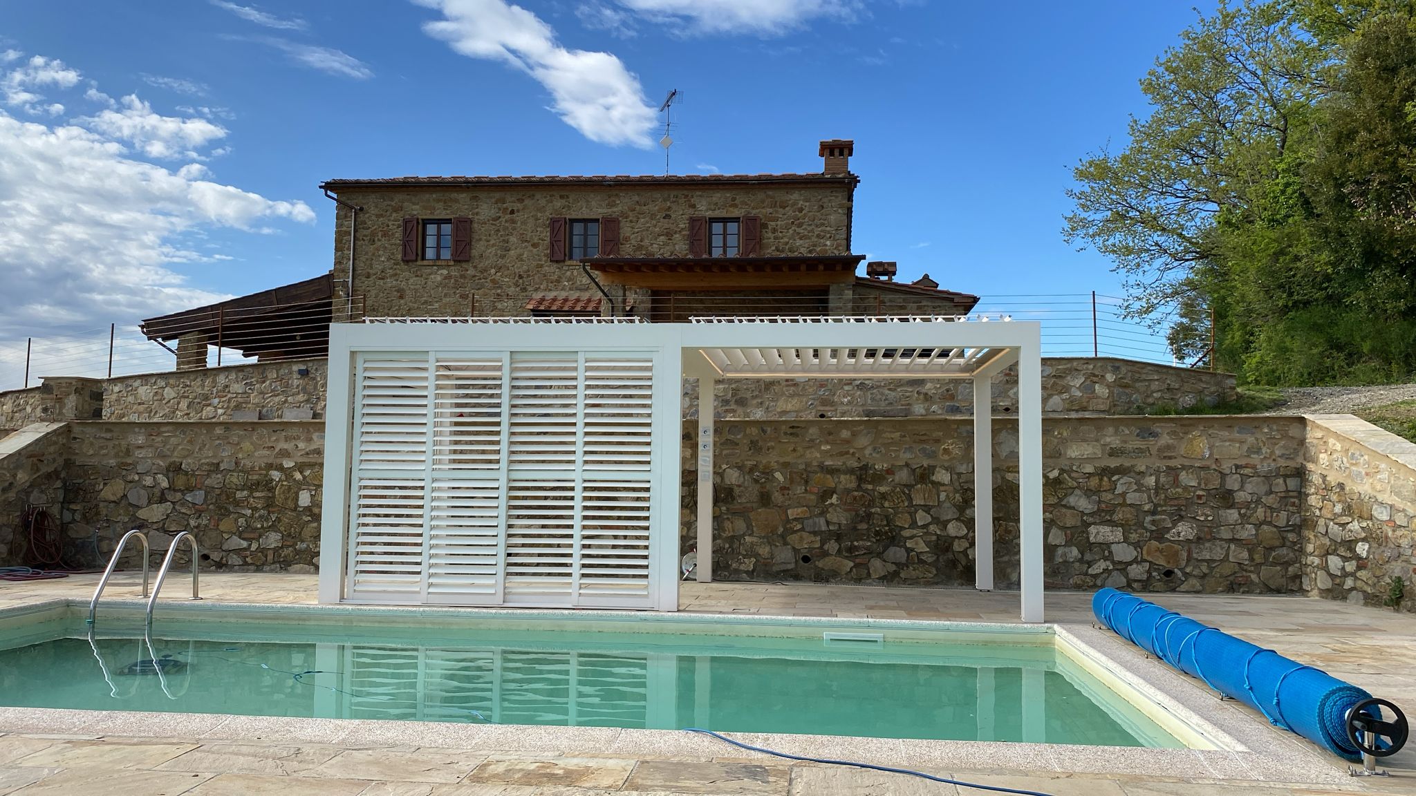 Weiße Pergola mit Lamellen-Schiebeelement am Pool vor einem Steinhaus