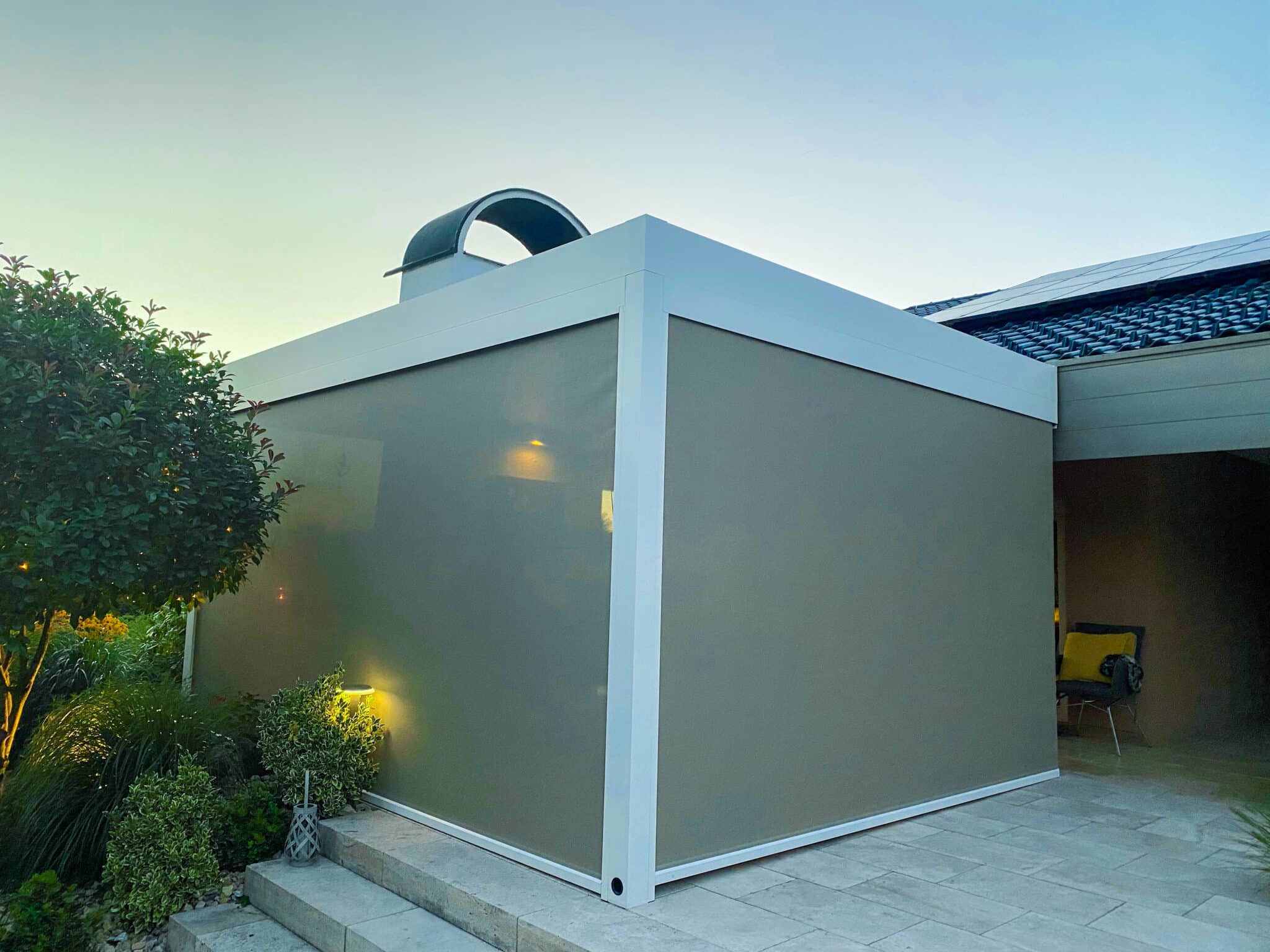 Weißes Lamellendach mit beigen ZIP-Screens in der Abenddämmerung mit Gartenbeleuchtung
