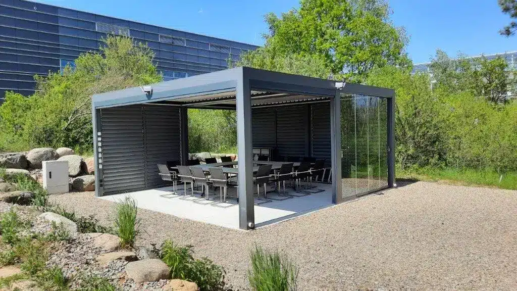Moderne Pergola mit Glaswänden und einem großen Tisch mit Stühlen im Inneren, umgeben von Grün. | my-pergola24.com