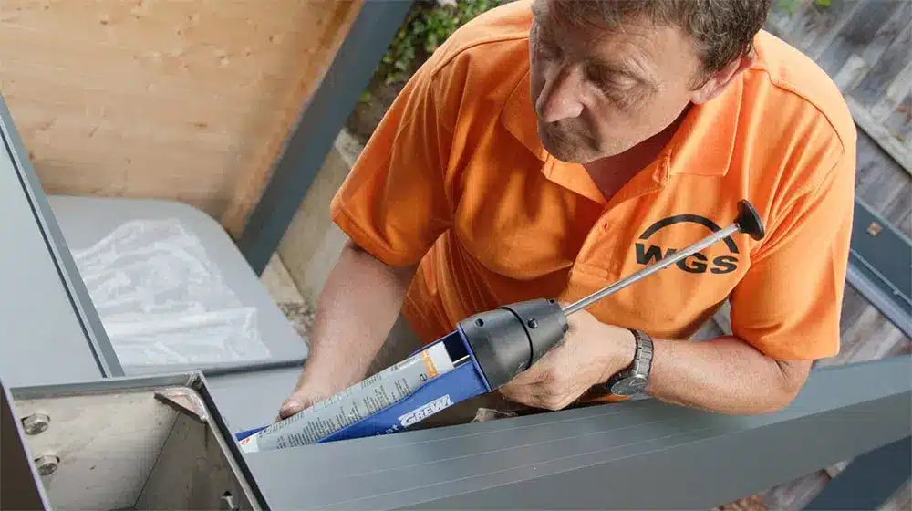Ein Mann in einem orangefarbenen Shirt verwendet eine Silikonpistole zur Montage eines Objekts im Freien. | my-pergola24.com