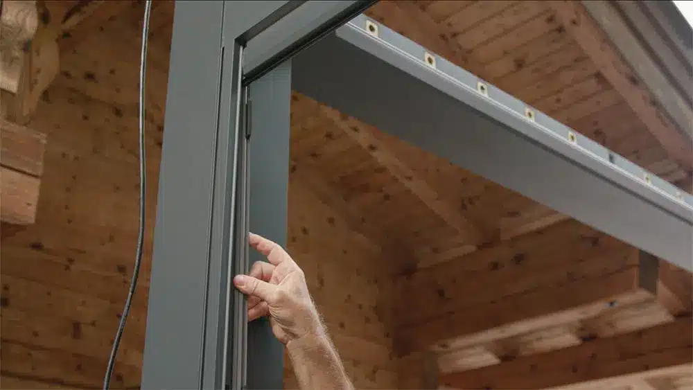Eine Hand, die eine Pergola-Komponente an einem grauen Rahmen anpasst, mit Holzstruktur im Hintergrund. | my-pergola24.com