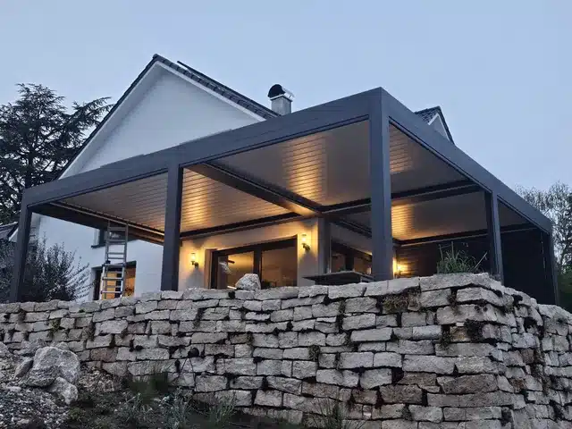 Lamellendach in Anthrazit am Abend, mit Licht in den Lamellen. Davor eine Mauer aus Naturstein