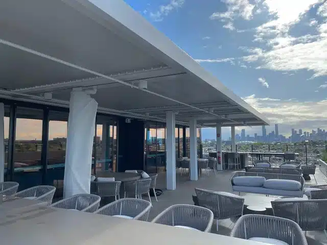 Großes weißes Lamellendach auf einer Dachterrasse. Dazu Lounge-Möbel und Blick auf die Stadt