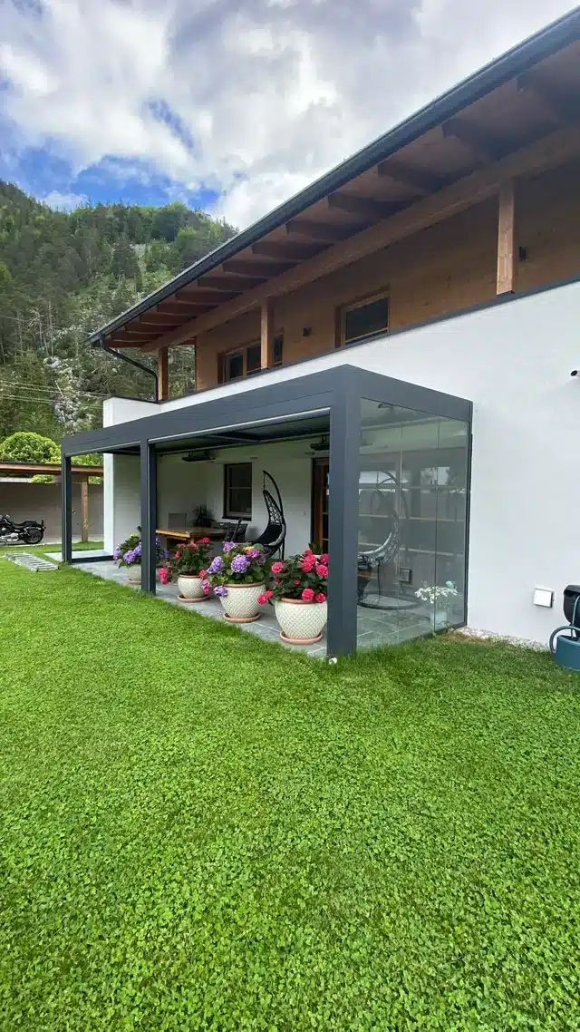 Lamellendach in Anthrazit mit Glasschiebewand an einem Haus in den Bergen, davor Hortensien in Töpfen