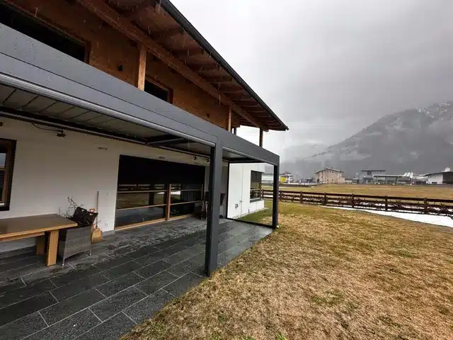 Ein Lamellendach in Anthrazit über der Terrasse. Hinten Berge bei Regen