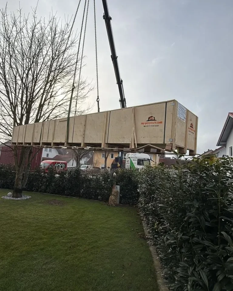 Kranwagen hebt die Holzkiste mit einem Lamellendach in den Garten