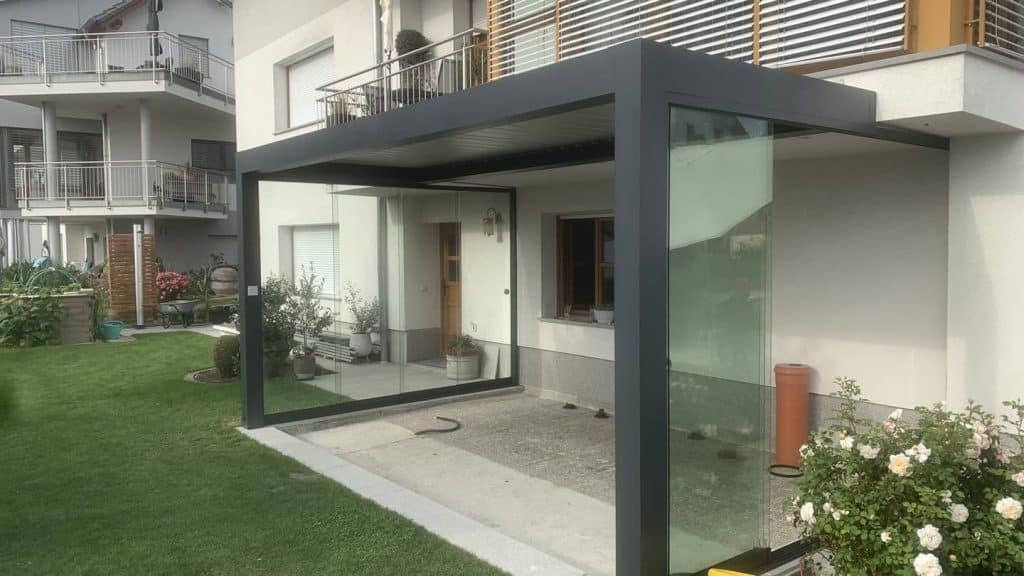 Anthrazitfarbene Pergola mit Glaswand am Erdgeschoss eines Mehrfamilienhauses, davor Rosen im Garten