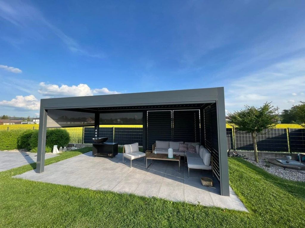 Anthrazitfarbene Pergola mit Loungeecke auf einer Steinterrasse im Garten, dahinter ein Rapsfeld