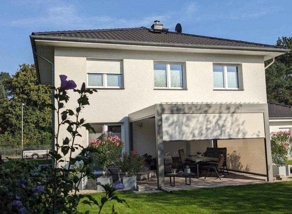 Beigefarbene Pergola in Falkensee mit Screen an einem Haus mit Walmdach, davor blühender Oleander