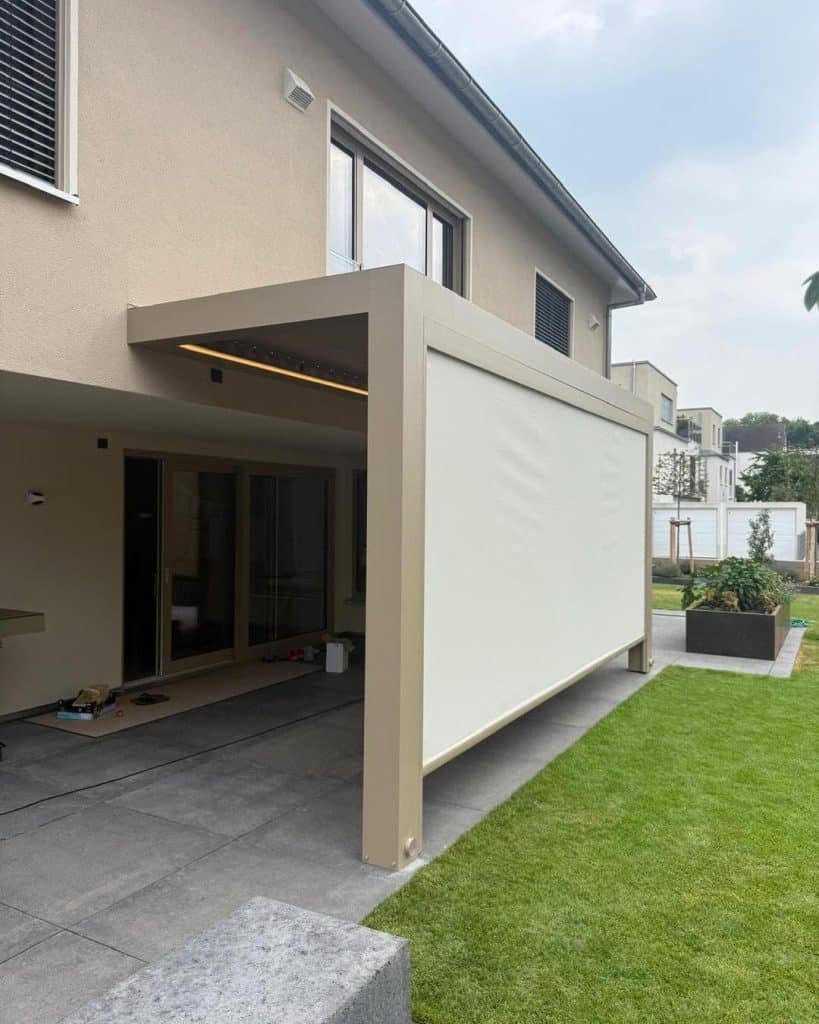Beige Pergola in Landau mit Seitenscreen unter dem Obergeschoss eines Hauses, davor Rasen und Terrassenplatten