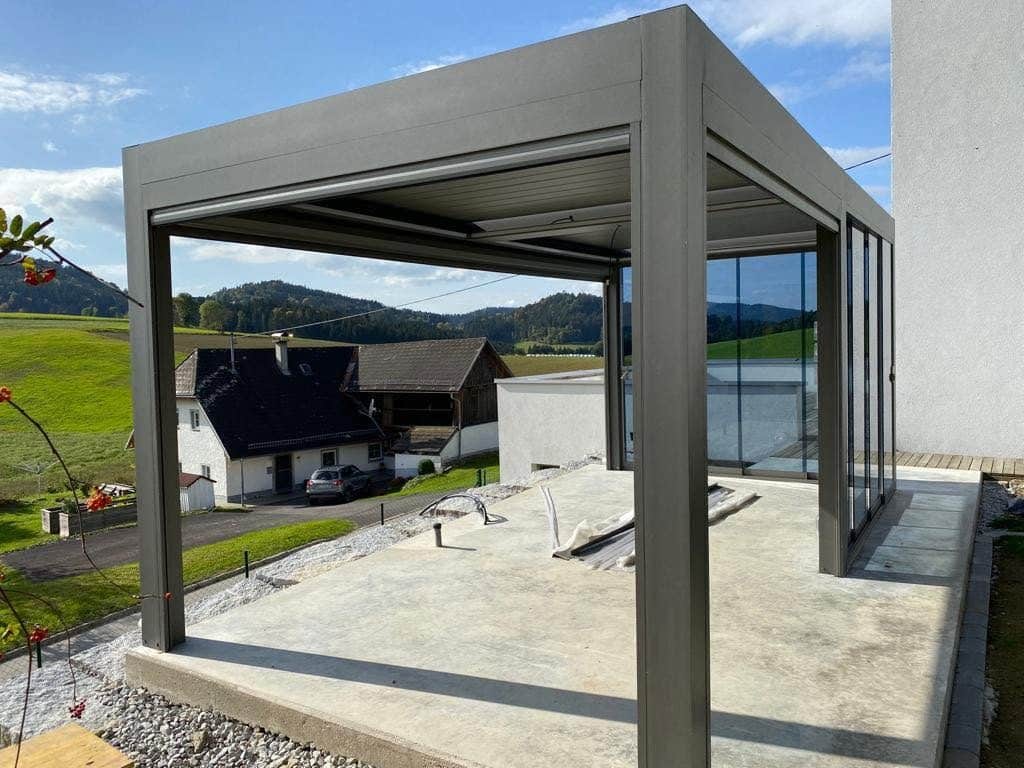 Anthrazitfarbene Pergola in Österreich auf einer Betonplatte, im Hintergrund Wiesen und Häuser