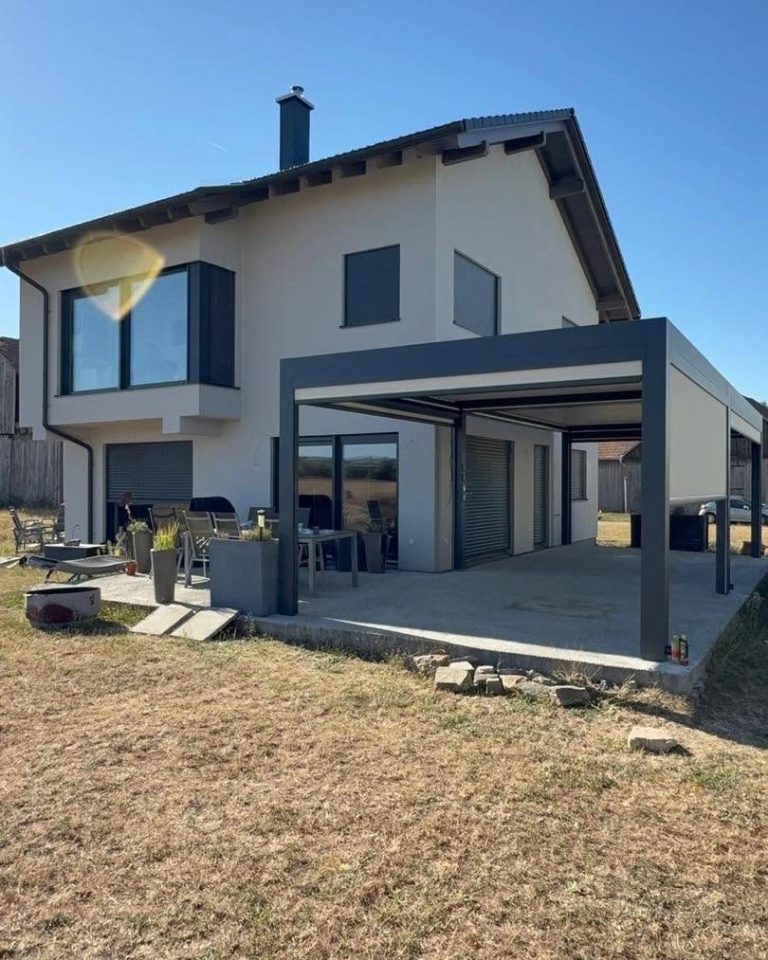 Pergola in Niederösterreich mit Lamellendach an einem Einfamilienhaus mit Erker, im Garten trockenes Gras