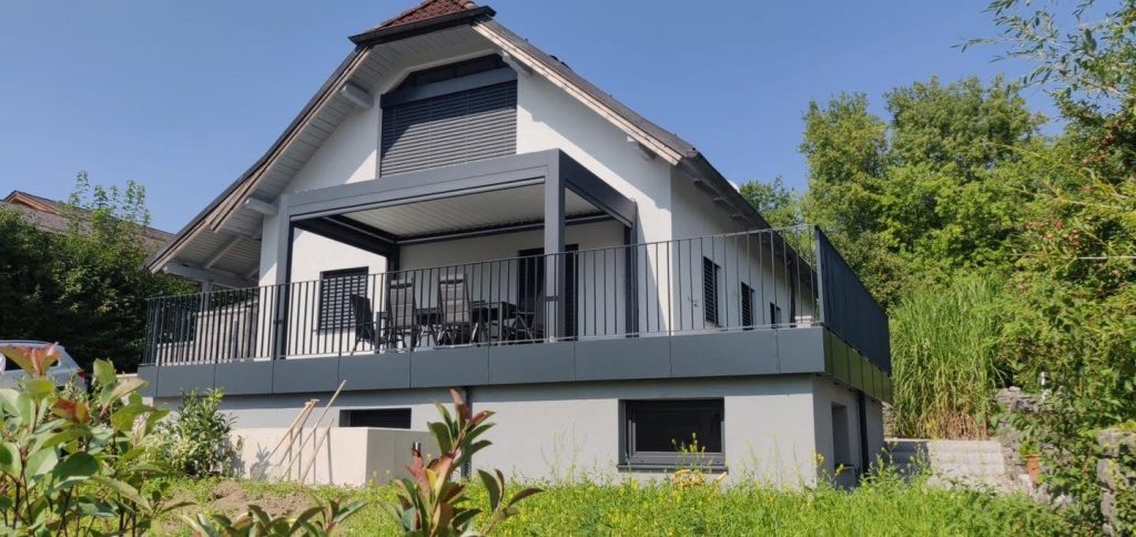 Pergola auf der erhöhten Terrasse eines Hauses mit Giebel, grauem Geländer und Natursteinmauer