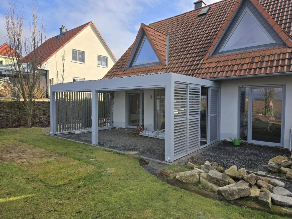 Fertiges graues Lamellendach mit Lamellenwänden am Haus mit rotem Ziegeldach, davor Natursteine im Beet