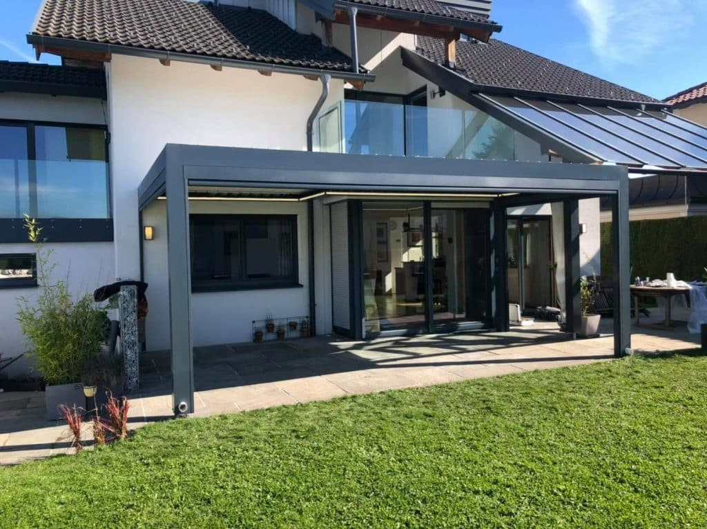 Anthrazitfarbene Pergola mit Glasgeländer darüber an einem Haus mit Giebel, davor ein Rasen im Sonnenschein