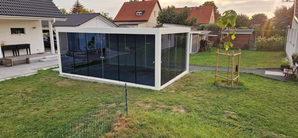 Glaspavillon mit Lamellendach mit anthrazitfarbenem Rahmen und dunklen Glaselementen im Garten, davor ein Rasen