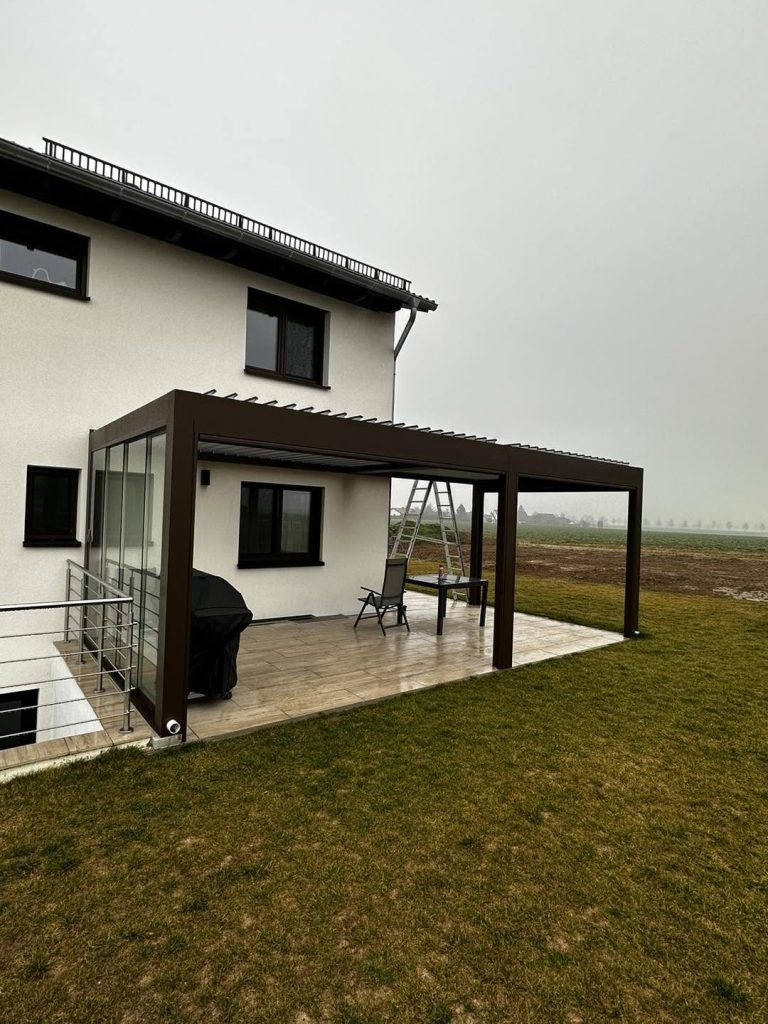 Pergola mit Glasschiebewand am Haus in Hanglage, davor ein Rasen