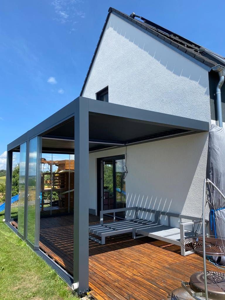 Anthrazitfarbene Pergola mit Glaswand am Giebelhaus, davor eine Holzterrasse und ein Sofa