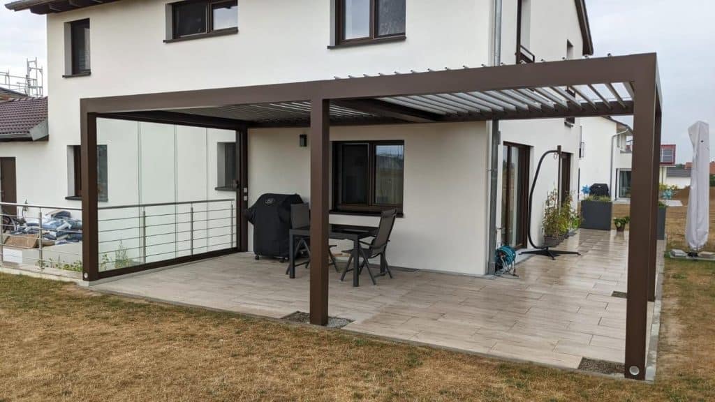 Braune Pergola mit Glaswand am Haus, auf der Terrasse ein Tisch und im Hintergrund ein Hängesessel
