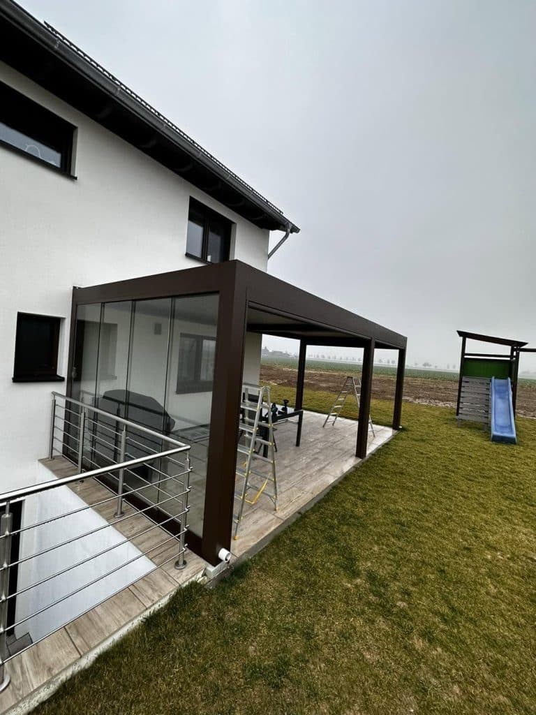 Braune Pergola mit Glaselement am Haus in Hanglage, daneben ein Edelstahlgeländer