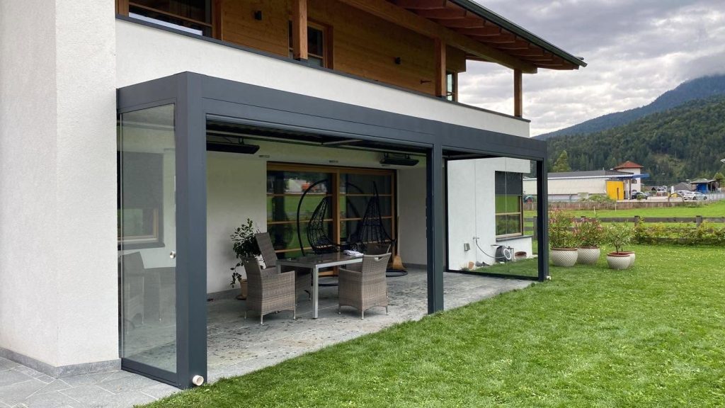 Anthrazitfarbene Pergola in Scharnitz mit Glaswand an einem Haus mit Holzbalkon, im Hintergrund Berge