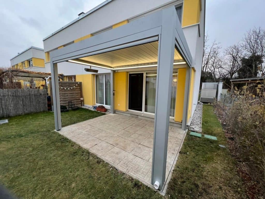 Graue Pergola mit LED-Licht am Reihenhaus mit gelber Fassade, davor Rasen und Natursteinplatten