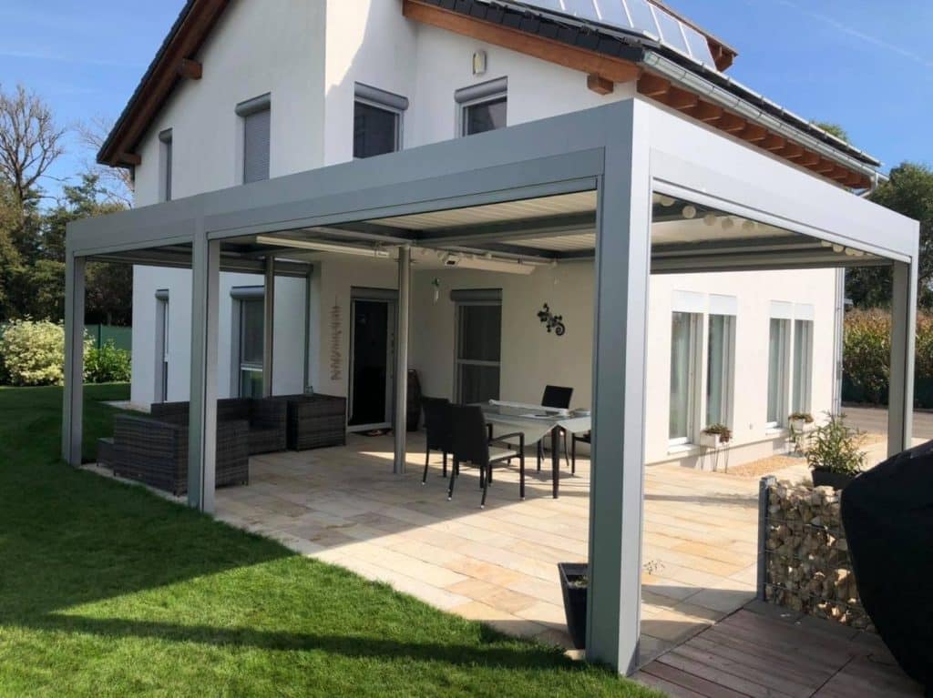 Graue Pergola über einer Terrasse mit Sitzmöbeln und einem Gabionen-Beet an einem Haus mit Solardach