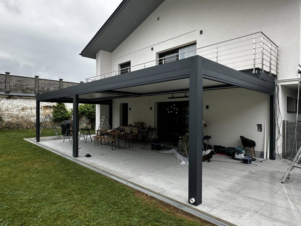 Große anthrazitfarbene Pergola in Völkermarkt über einer Terrasse an einem Haus mit Balkon und Edelstahlgeländer