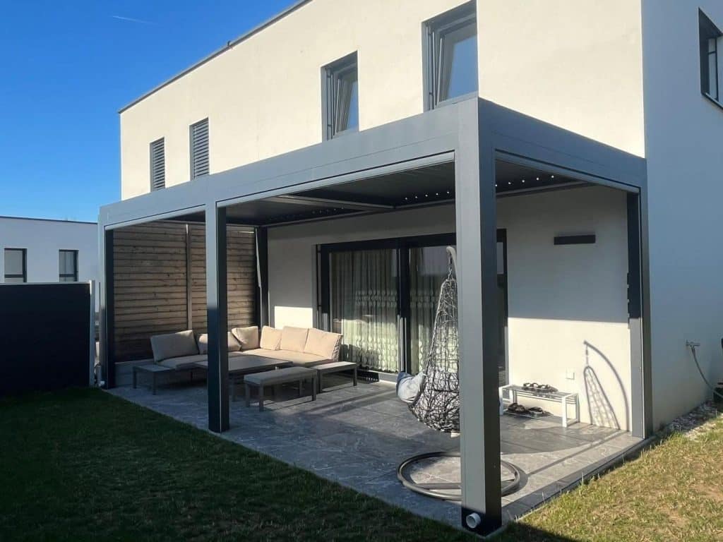 Pergola mit Hängesessel und Sofa auf einer Terrasse am Haus, davor ein Rasen