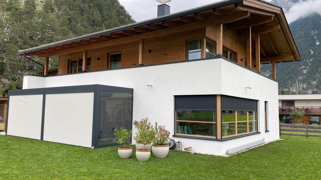 Haus mit Holzbalkon und Pergola in Scharnitz, an der Seite eine Lamellenwand mit Glas, im Hintergrund Berge
