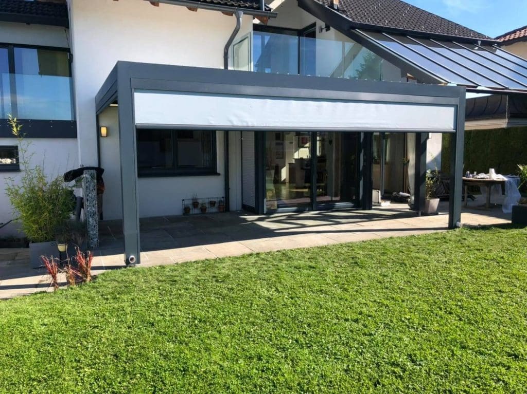 Anthrazitfarbene Pergola mit hellem Screen über der Terrasse eines Hauses, davor ein gepflegter Rasen
