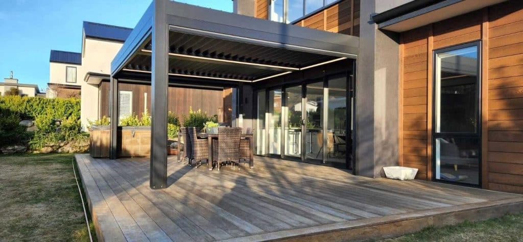 Anthrazitfarbene Pergola mit Holzterrasse an einem Haus mit Holzverkleidung, im Garten ein Rasen