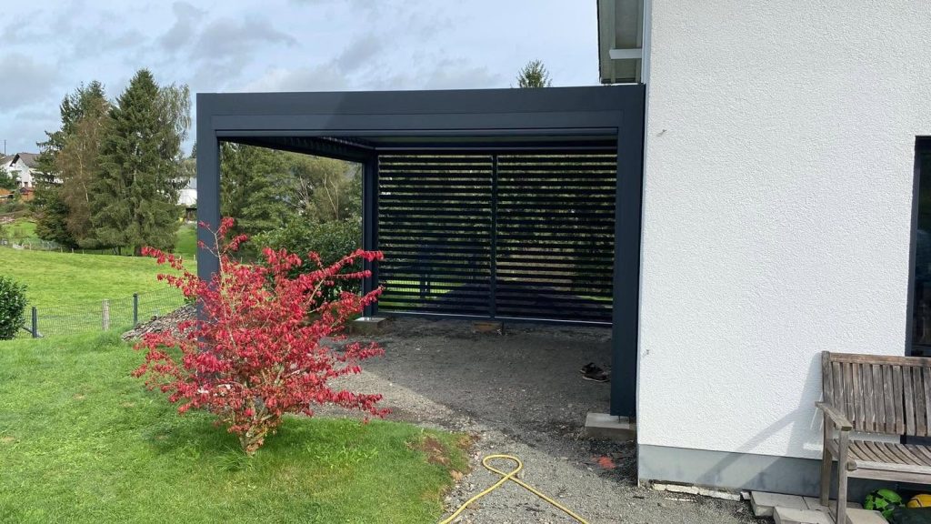Pergola in Siegen mit Lamellenwand am Haus, davor ein rot blühender Zierstrauch im Garten