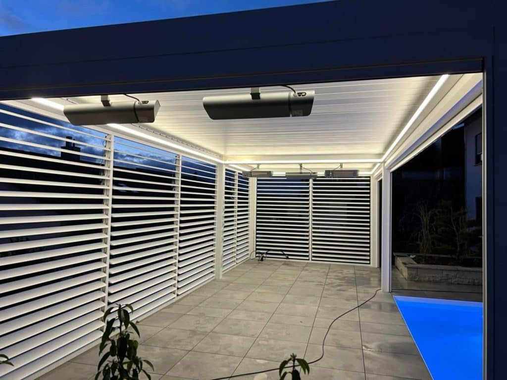 Weiße Lamellenwand unter einer Pergola in Reutlingen mit zwei Heizstrahlern und LED-Streifen, rechts der blau beleuchtete Pool