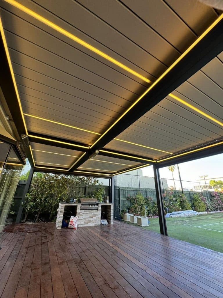 Lamellendach mit LED-Streifen über einem Holzdeck, im Hintergrund eine Outdoorküche