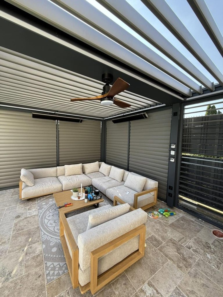 Loungeecke mit hellem Sofa unter einem Lamellendach in Braunau mit Deckenventilator und Lamellenwand