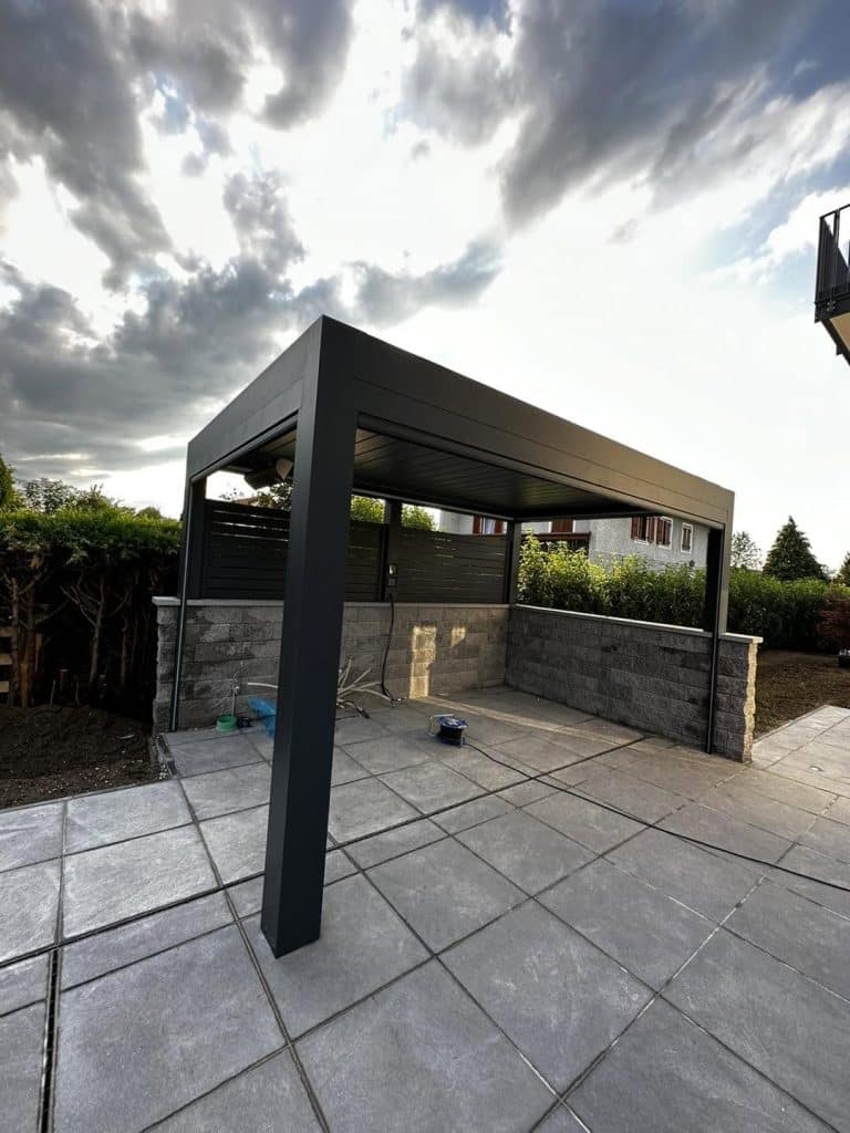 Anthrazitfarbene Pergola mit Natursteinmauer und Grillplatz im Hintergrund, bewölkter Himmel