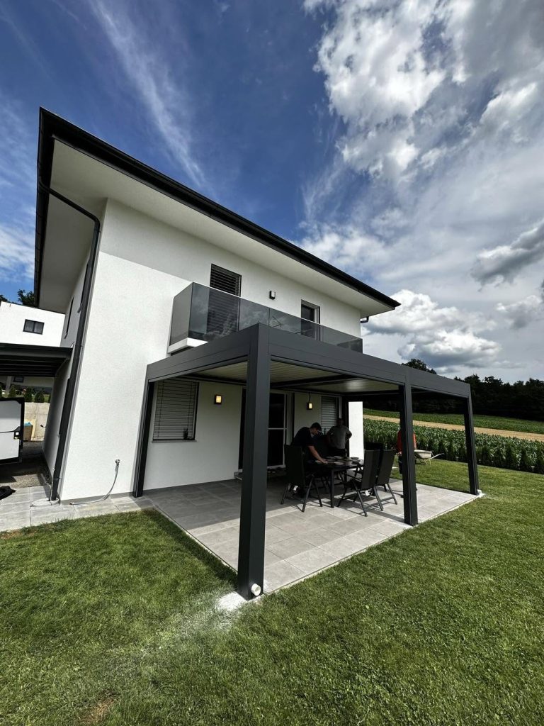 Anthrazitfarbene Pergola an einem Neubau, davor ein frisch gemähter Rasen, bewölkter Himmel