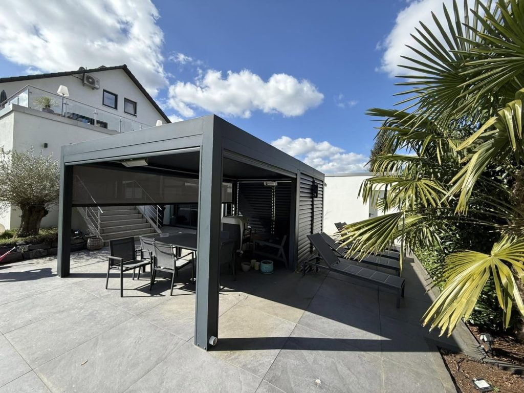 Lamellendach mit Gartenmöbeln und Palme auf einer Terrasse mit Steinplatten, dahinter ein Wohnhaus