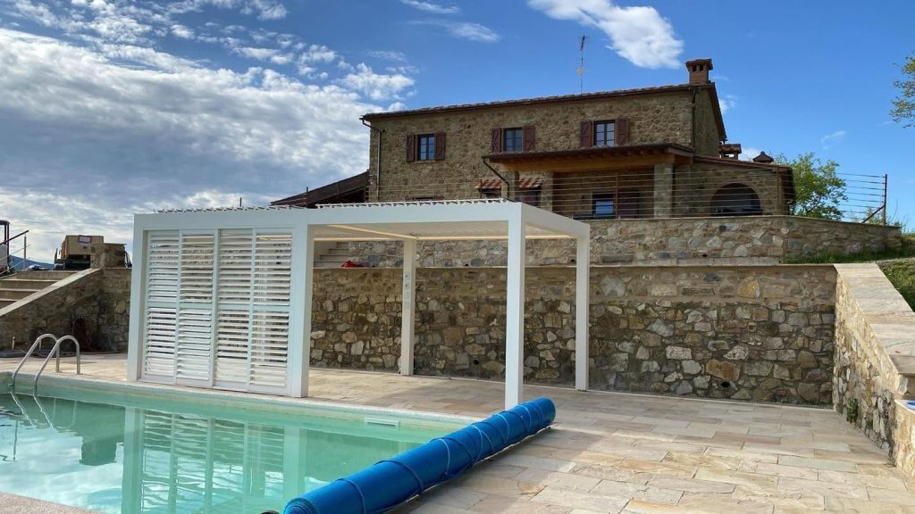 Pool mit weißem Lamellendach in der Toskana und Lamellenwänden, dahinter ein Haus aus Naturstein