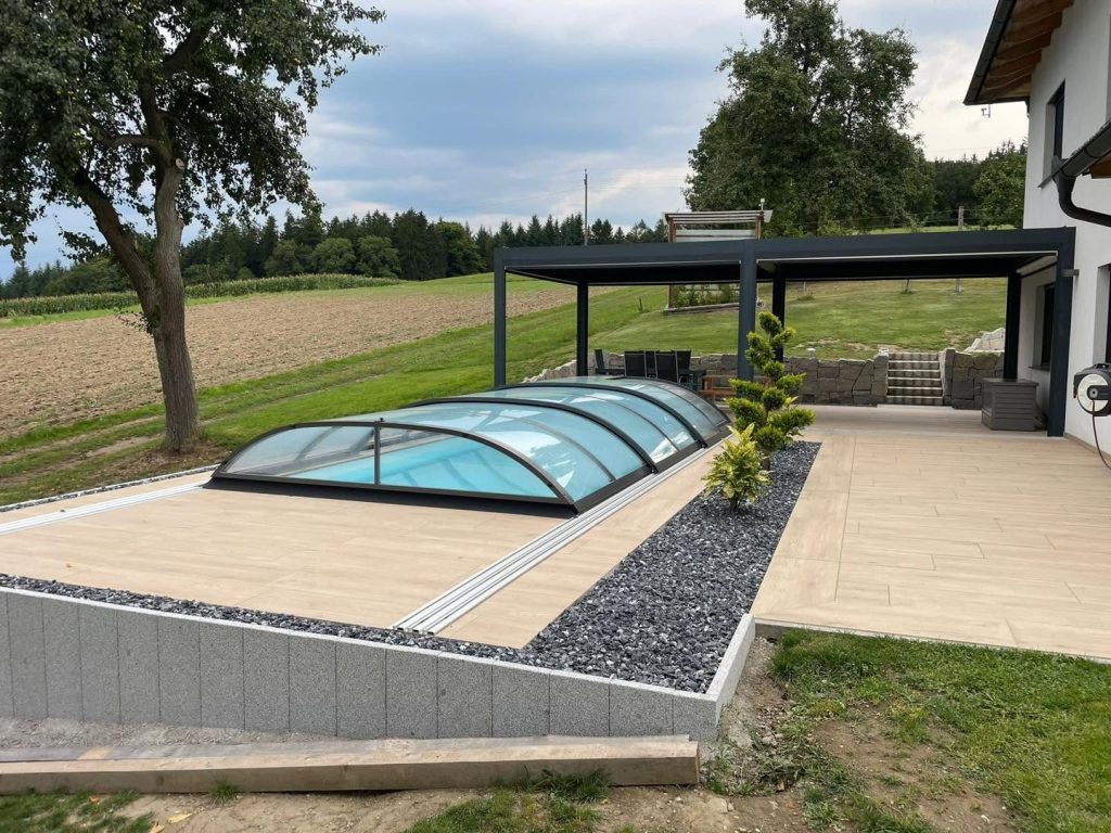 Pergola auf einer Terrasse mit Poolüberdachung, dahinter Felder und ein Haus