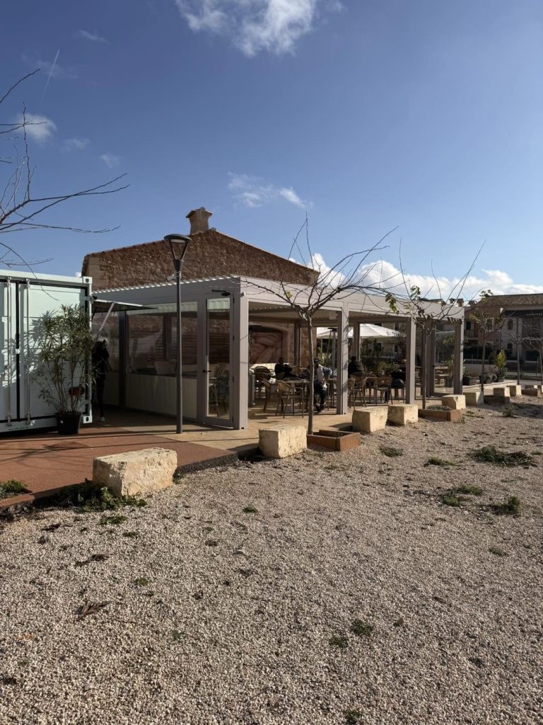 Restaurant mit Lamellendach-Terrasse vor einem Steinhaus, davor eine Kiesfläche