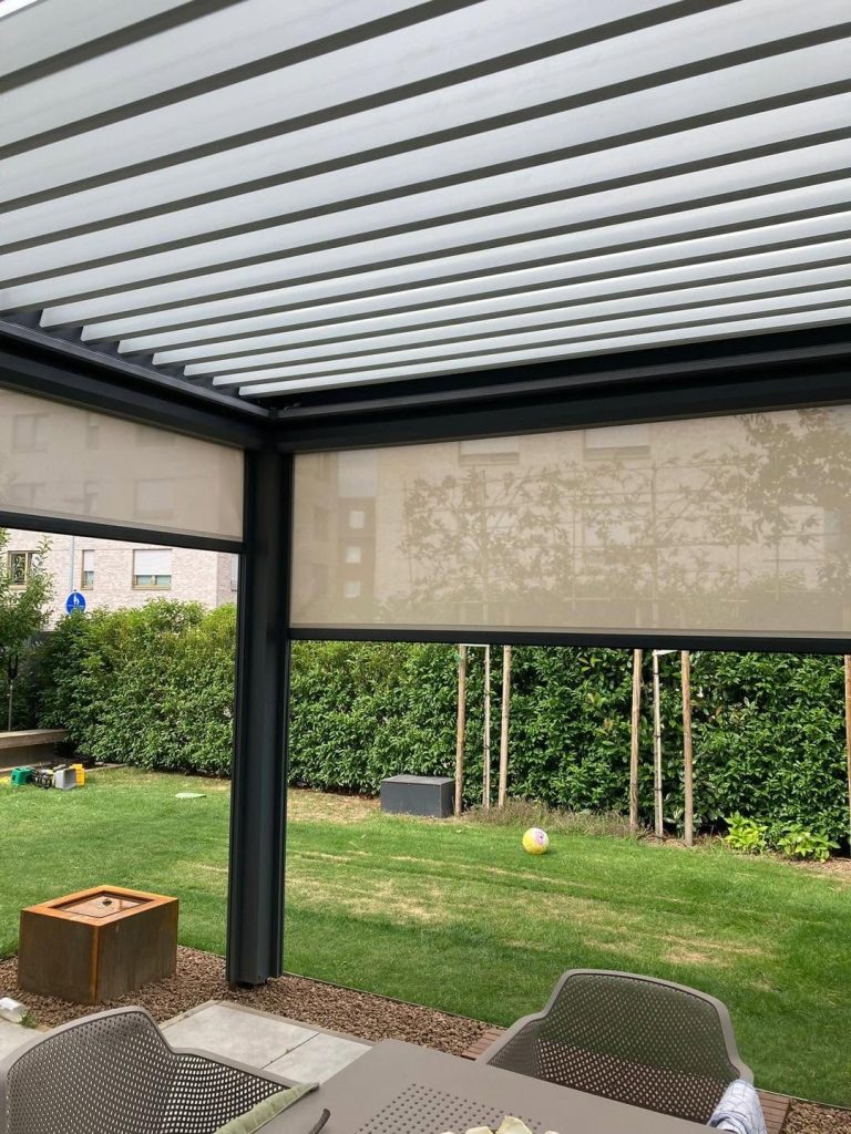 Lamellendach mit Screen unter geöffneten Lamellen, dahinter ein Garten mit Hecke und Rasen