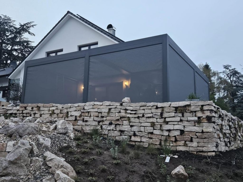 Lamellendach in Köln mit Screens über einer Trockenmauer aus Naturstein am Hang, die Beleuchtung brennt