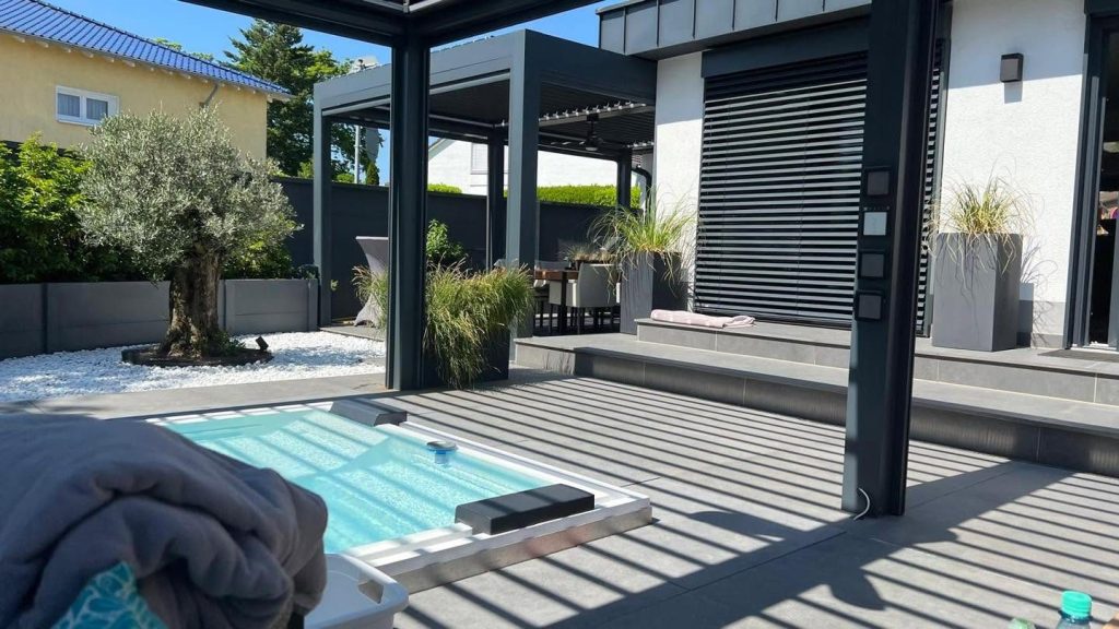 Pergola in Köln über einer Terrasse mit kleinem Pool und Olivenbaum, im Hintergrund ein Haus mit Rollladen