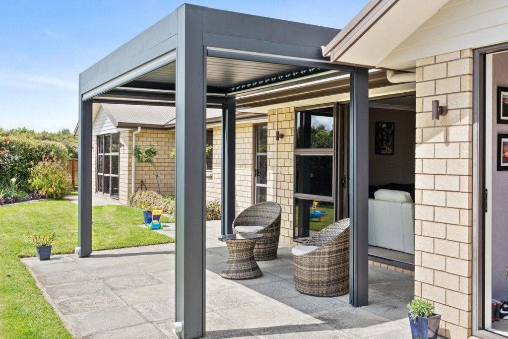 Anthrazitfarbene Pergola auf einer Terrasse mit Rattansesseln, daneben ein Backsteinhaus im Sonnenschein