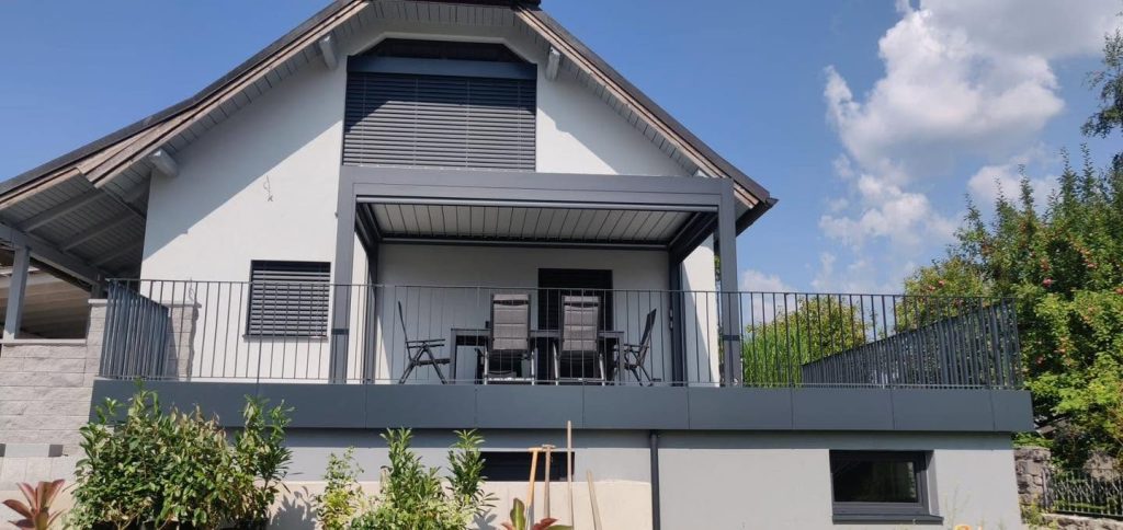Pergola mit Gartenmöbeln auf der erhöhten Terrasse eines Hauses, davor ein gepflegter Garten