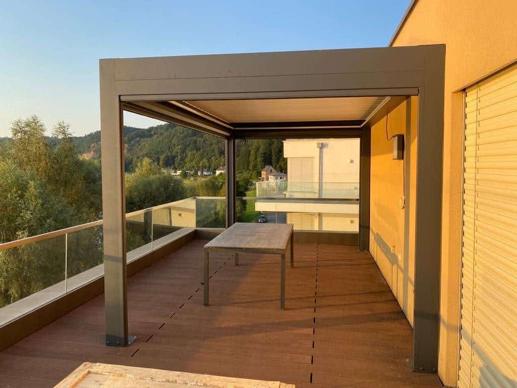Anthrazitfarbene Pergola mit Tisch auf einer Dachterrasse, links ein Glasgeländer, rechts Rollläden