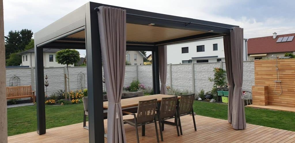 Anthrazitfarbene Pergola in Klagenfurt mit beigen Vorhängen und langem Esstisch auf einer Holzterrasse